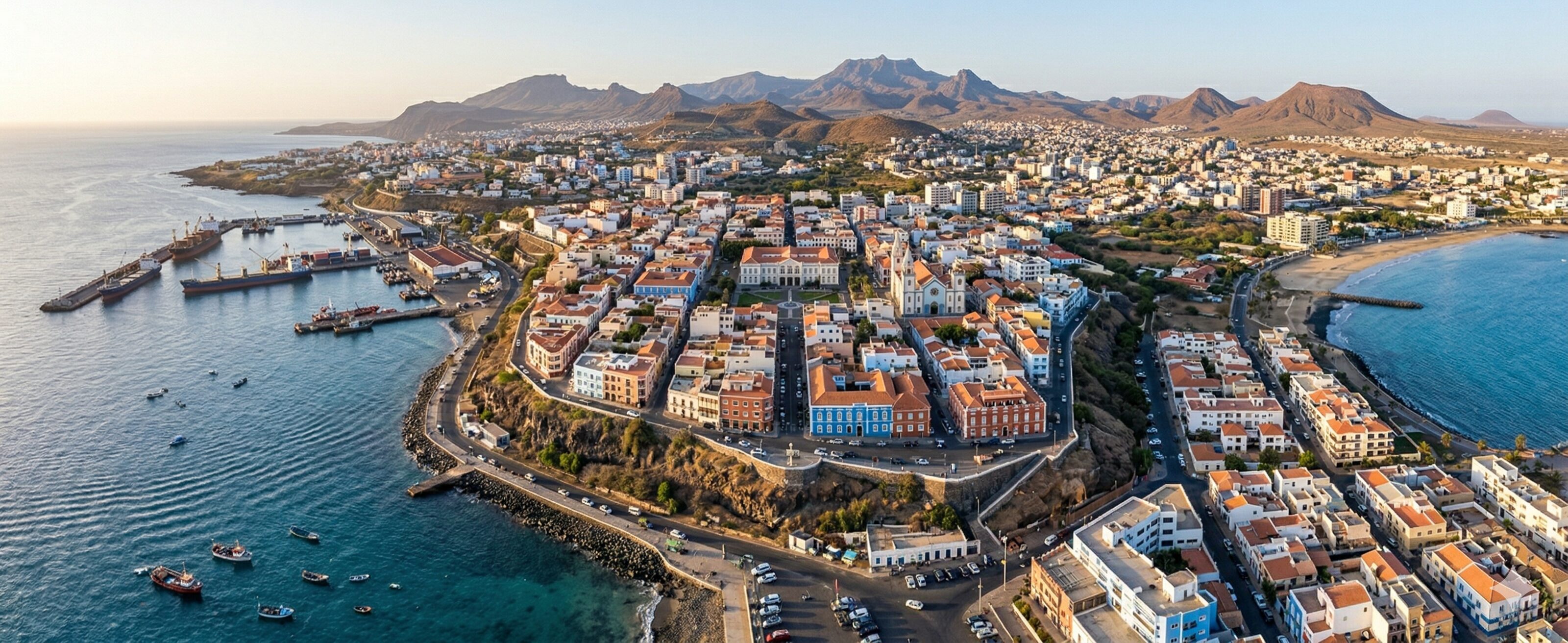 Stunning panoramic bird's-eye view of Praia, Cape Verde, at golden hour, showing the complex coastline with cliffs, a busy port, the historic Plateau district, and distant volcanic mountains.